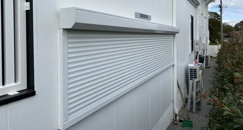 White roller shutter fitted to an exterior wall beside an air conditioning unit and narrow side path