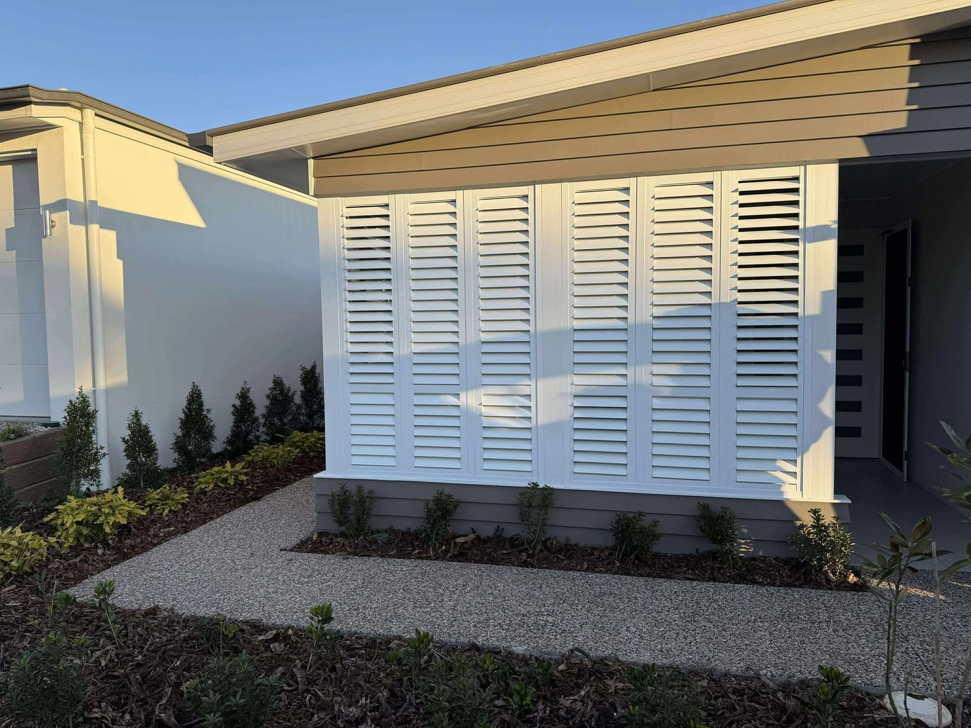 House exterior corner with cream plantation shutters enclosing the alfresco, garden bed alongside