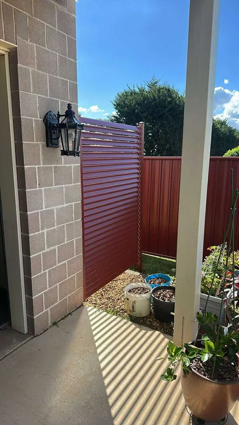 Red aluminium slat privacy screen enclosing a small paved courtyard, block wall and potted plants alongside
