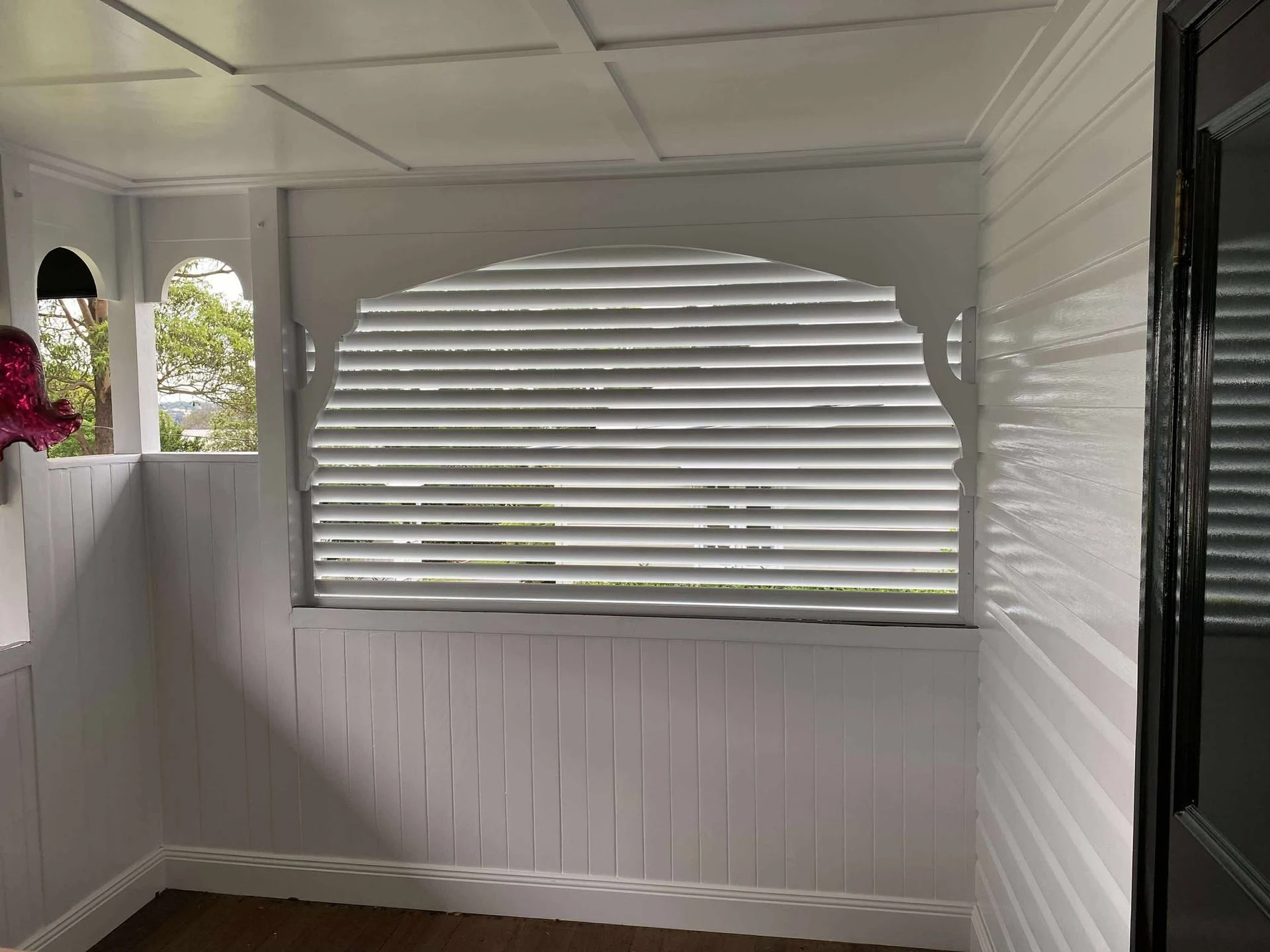 Arched verandah window fitted with a white louvre blind, lined timber walls around it